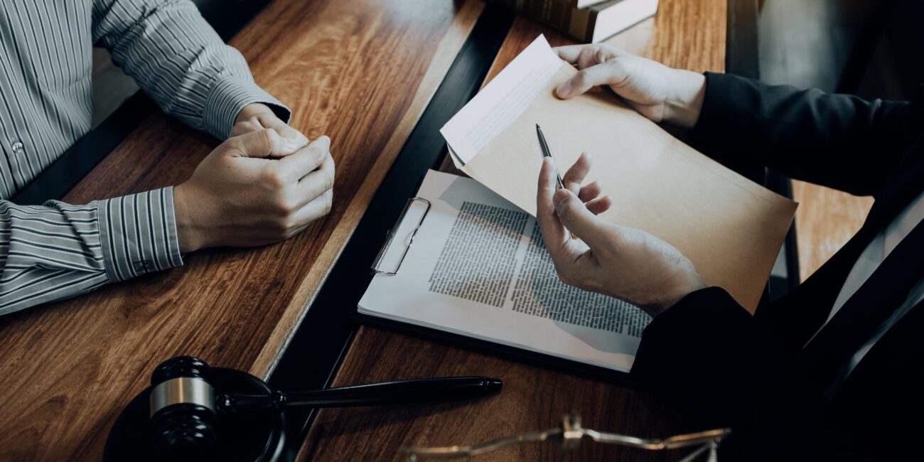 Primo piano di un incontro tra due persone in ufficio legale, con documenti su una cartellina, una penna e un martelletto giudiziario sul tavolo.