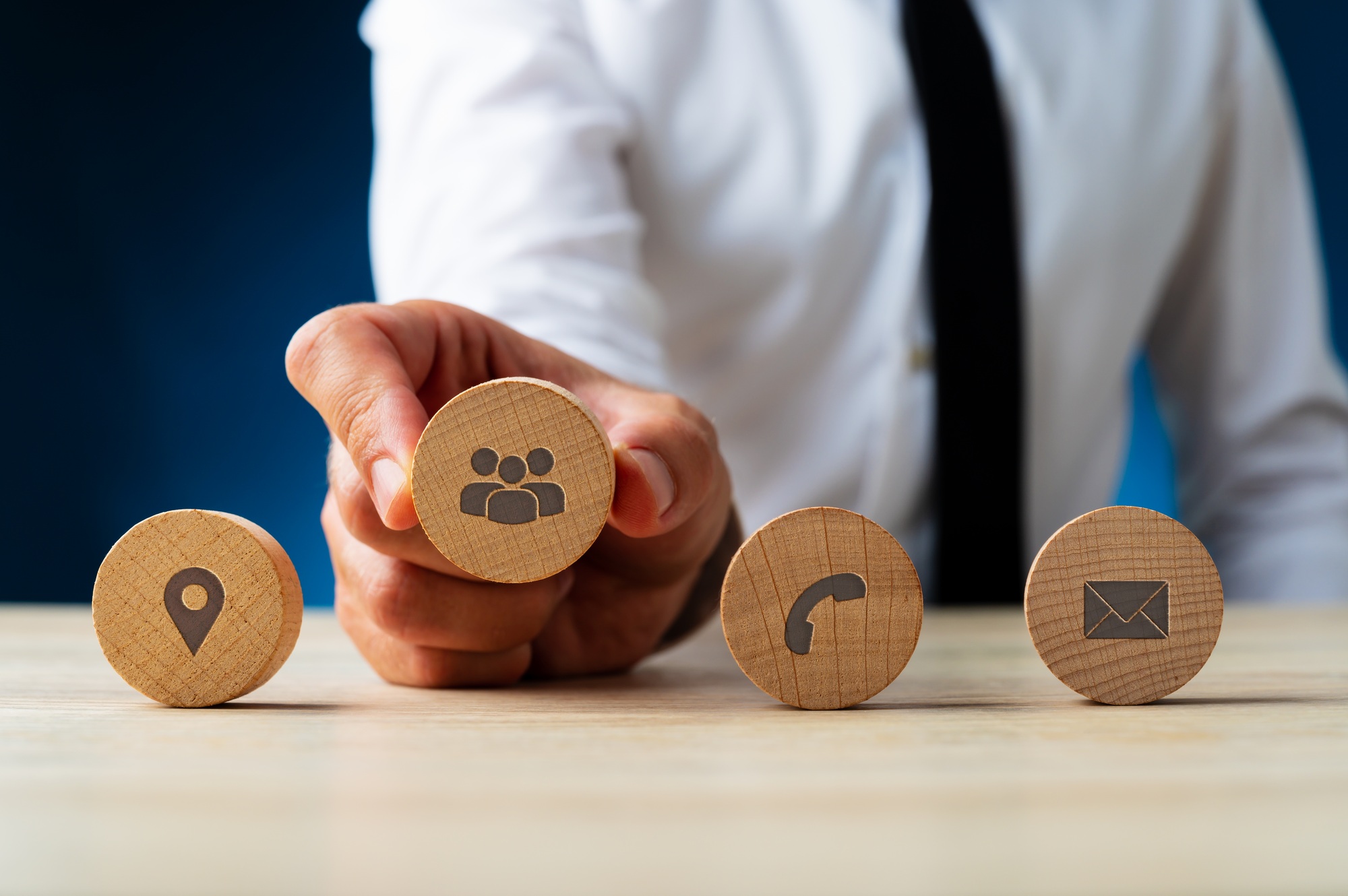 Persona in camicia e cravatta che tiene un disco di legno con icona di gruppo; accanto simboli di posizione, telefono ed email.